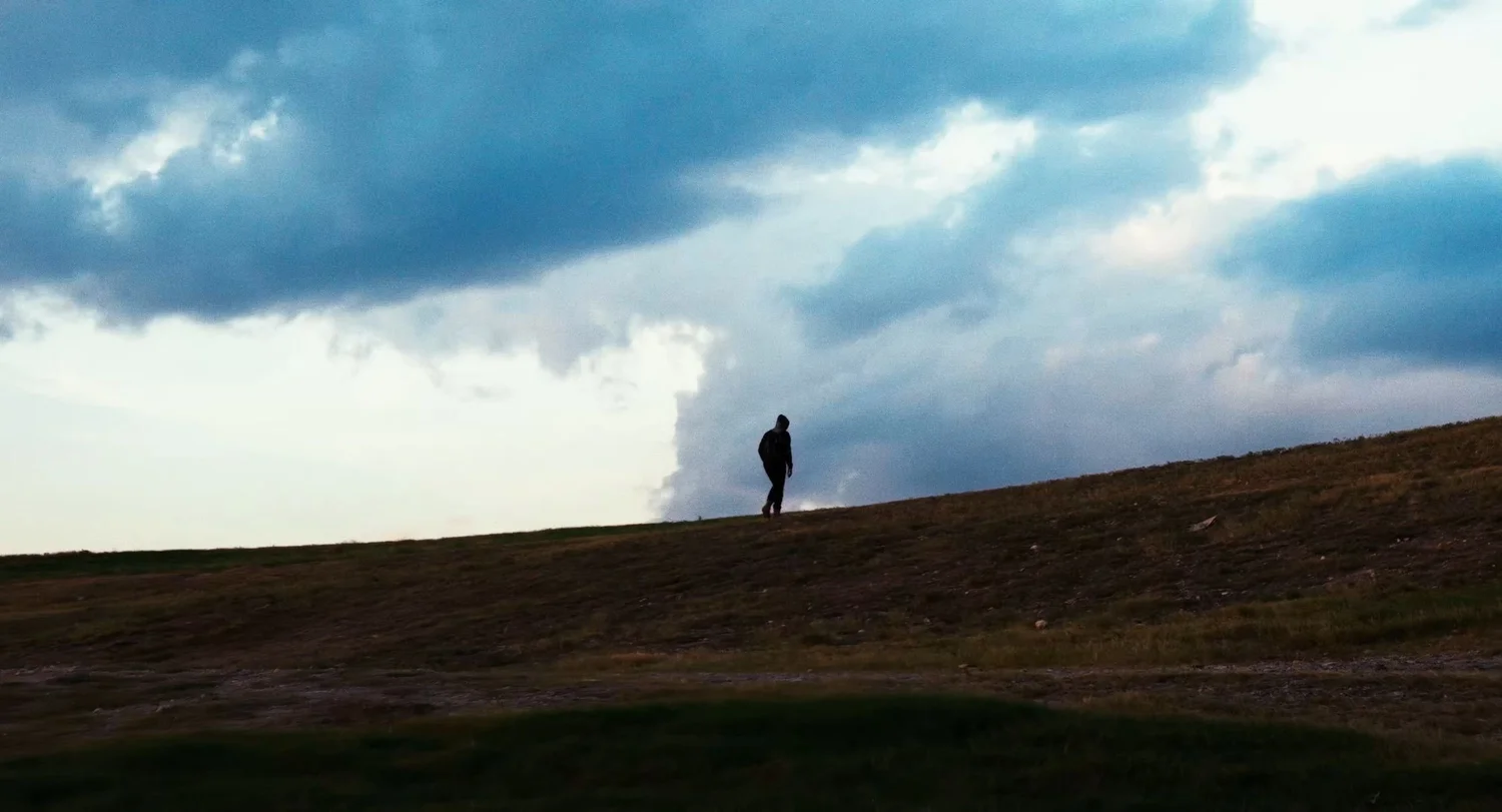 Still from INERTIA: a hooded figure in silhouette walks along the crest of a grassy hill under heavy clouds.