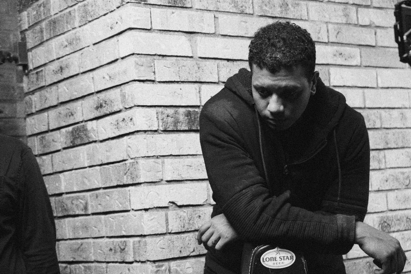 Behind the scenes on INERTIA: an actor in a black hoodie leans on a stand against a brick wall, looking down.