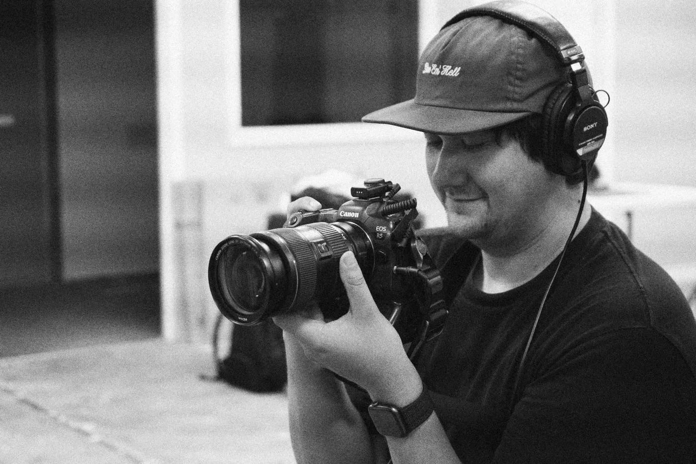 Behind the scenes on INERTIA: a smiling camera operator in a brown cap and headphones frames a shot with a long lens.