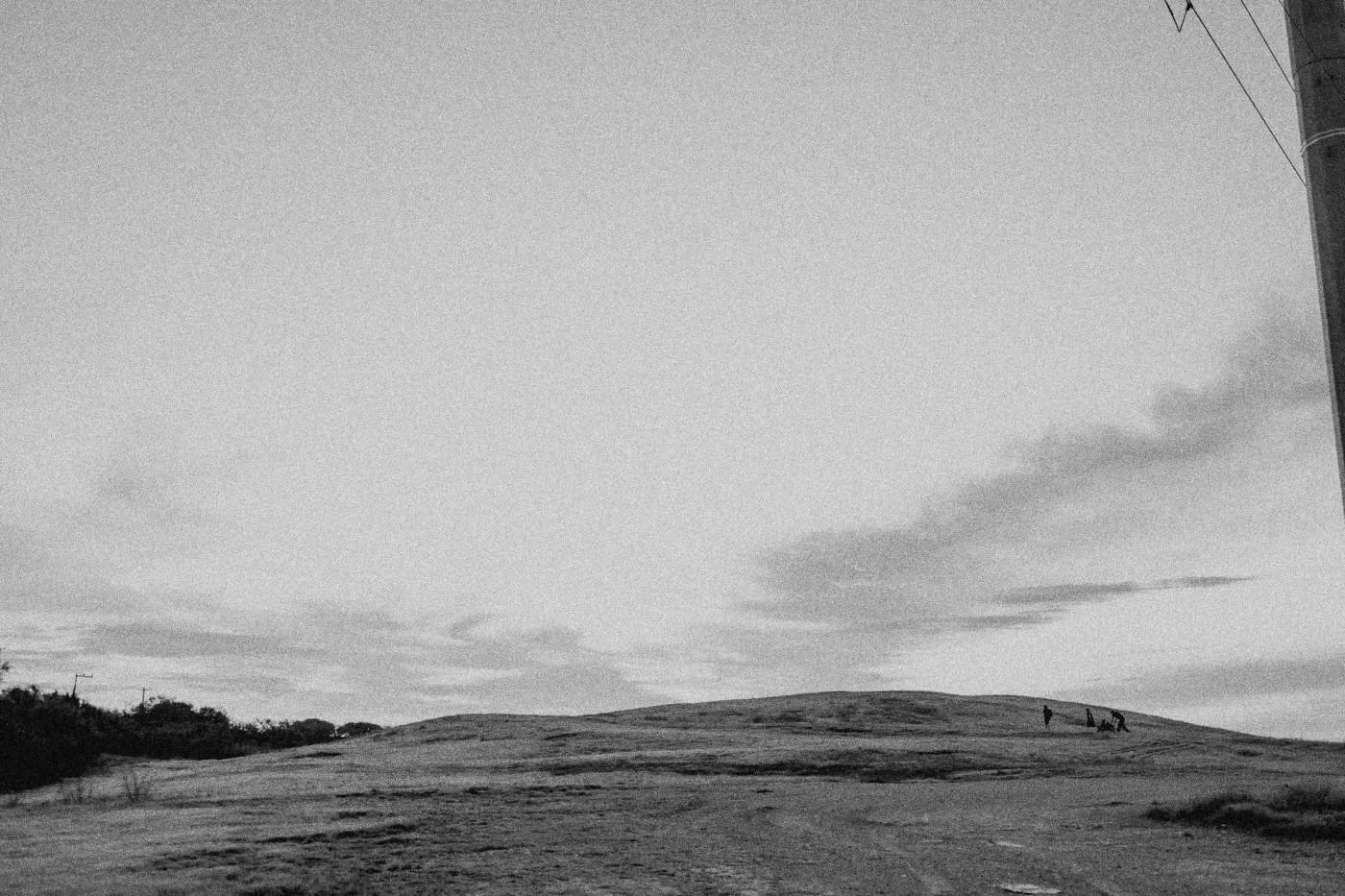 Behind the scenes on INERTIA: a wide hilltop at dawn under a pale sky, tiny figures on the ridge.