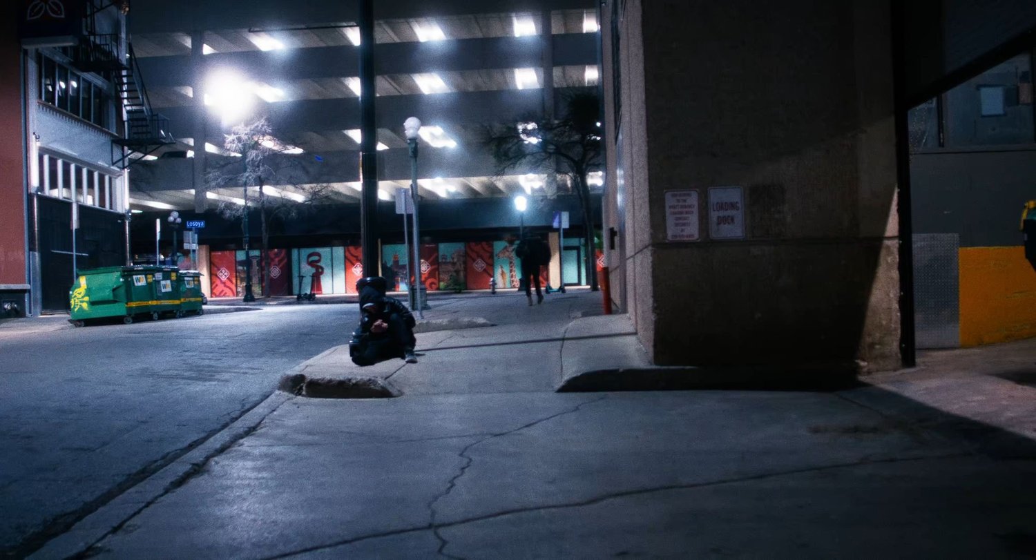 Still from INERTIA: a person in a dark hooded jacket crouches on a curb at night beneath a brightly lit parking garage while another person walks in the distance past painted murals.
