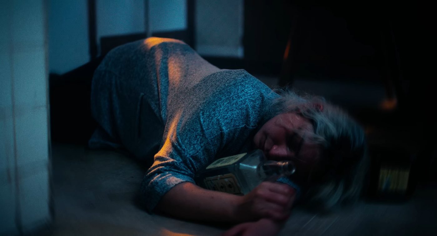 Still from INERTIA: a woman with platinum blonde hair lies curled on a floor holding a glass liquor bottle against her chest, in dim blue and amber light.