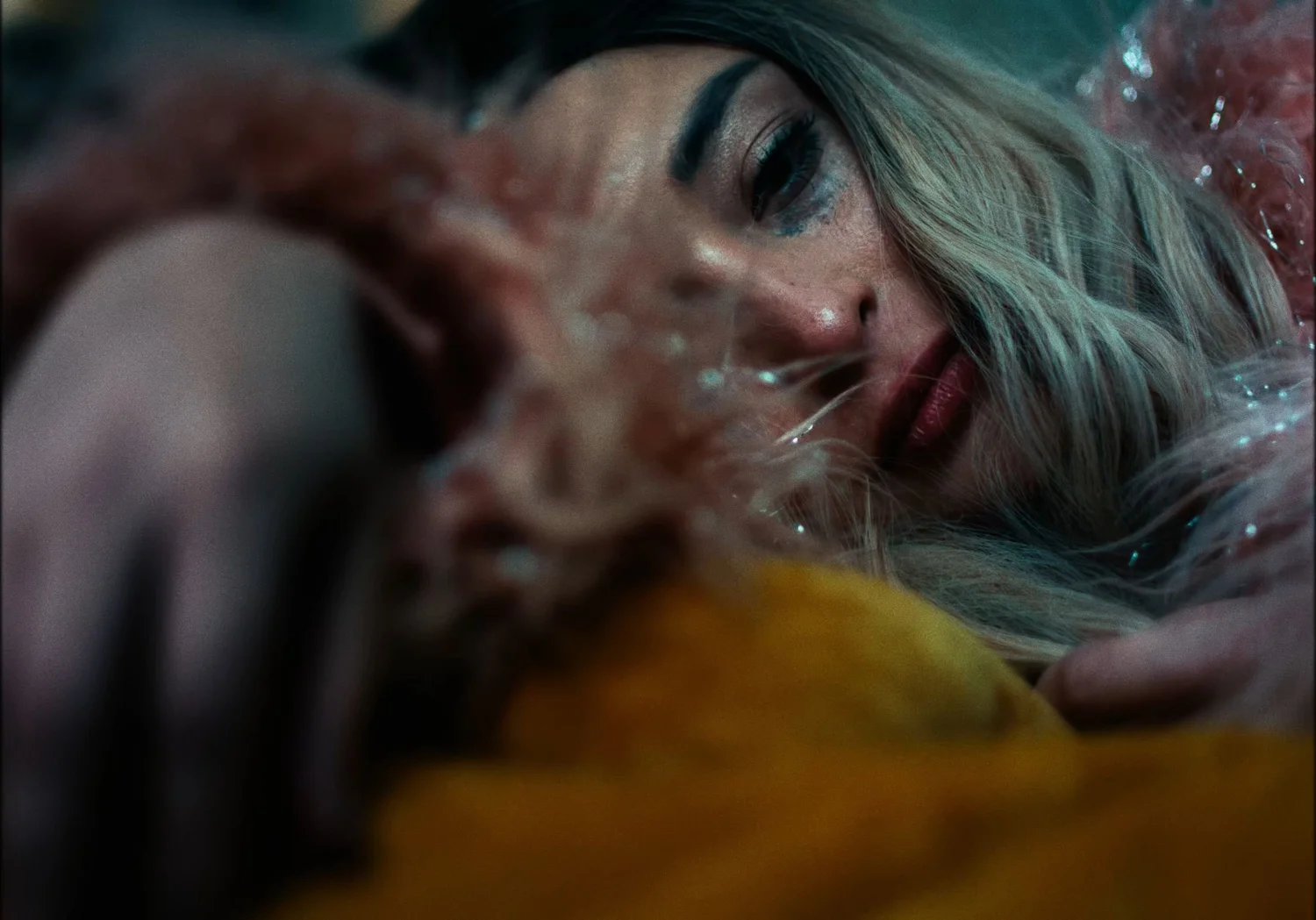 Still from INERTIA: a woman with blonde hair and smudged eye makeup lies on her side against mustard colored fabric, a pink feathered coat around her.