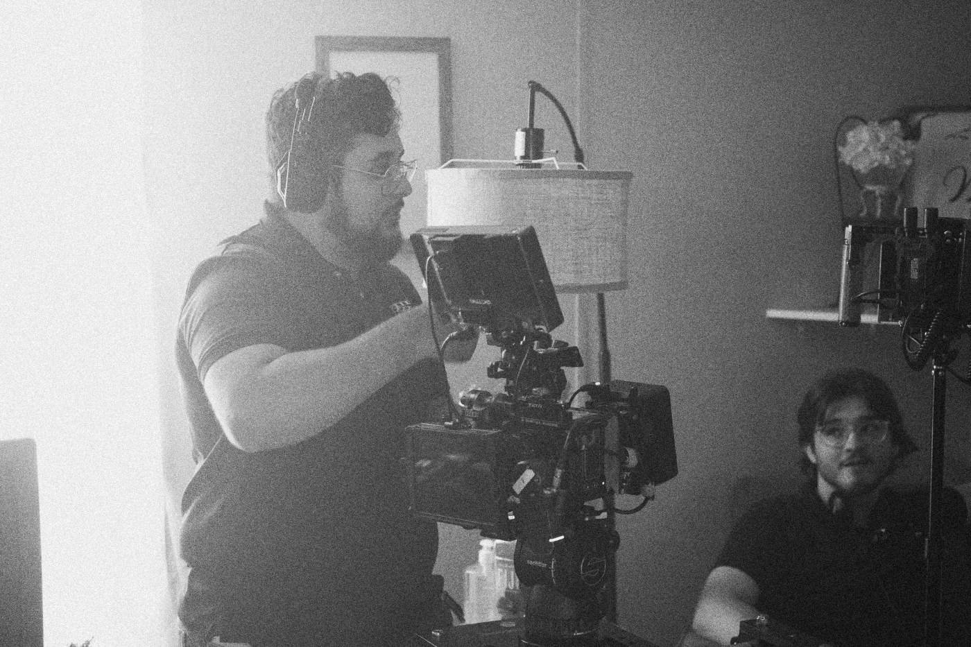 Behind the scenes on INERTIA: a camera operator in a headset works a rigged camera by a sunlit window, a colleague seated behind.