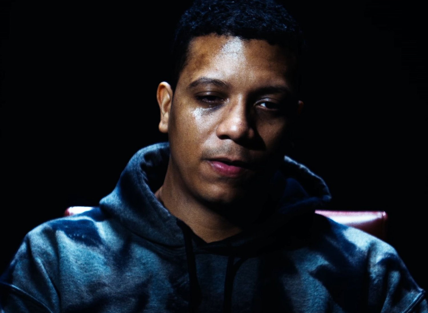 Still from INERTIA: a man in a dark hooded sweatshirt sits in a red chair against a black background, his face lit from above.