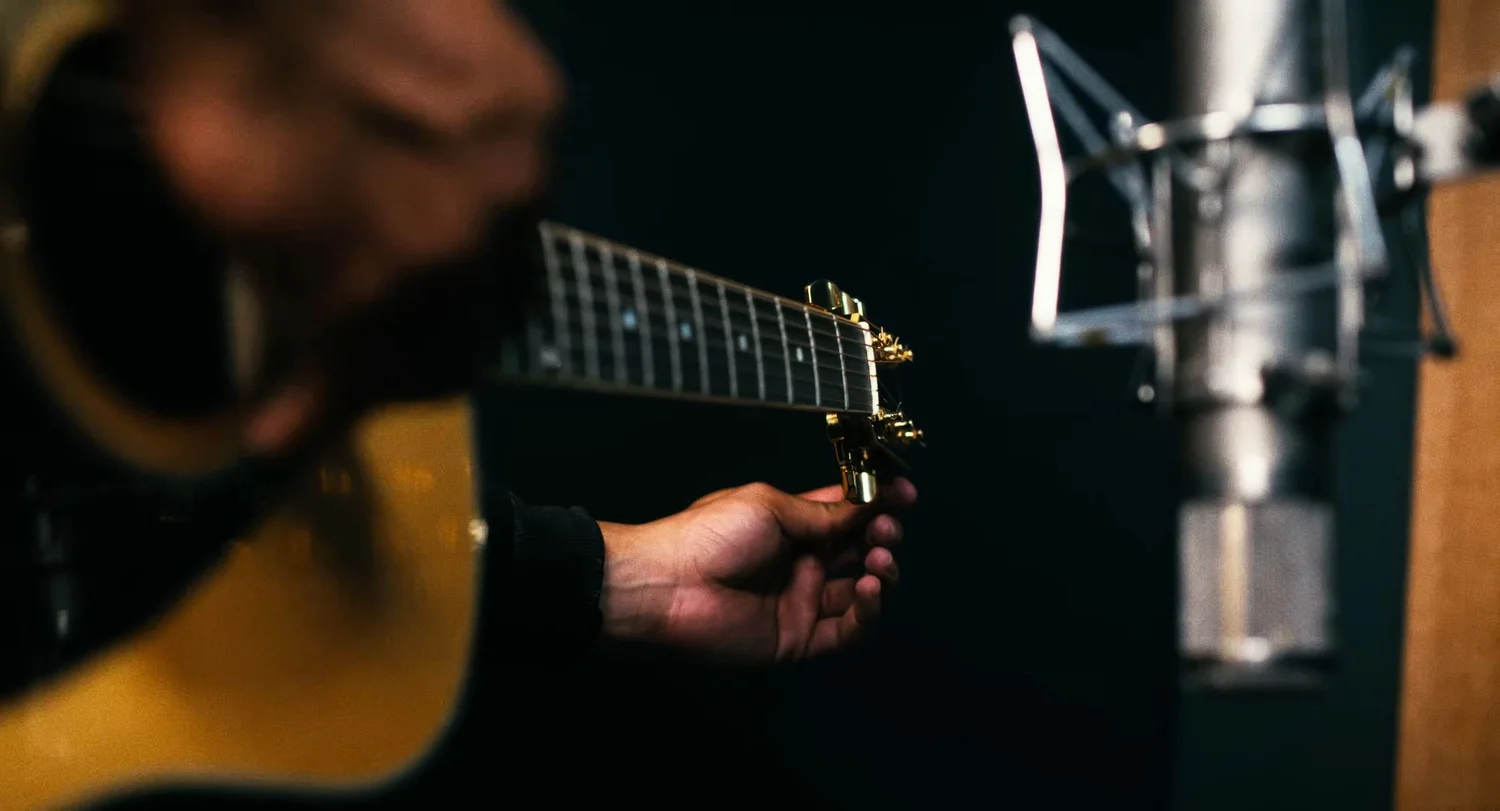 Still from INERTIA: a hand reaches for the tuning pegs of an acoustic guitar near a studio condenser microphone.