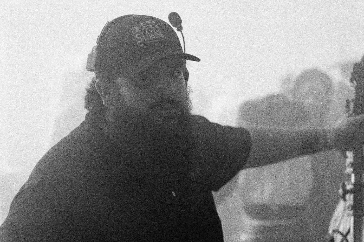 Behind the scenes on INERTIA: a bearded crew member in a cap and headset looks toward the camera through haze.