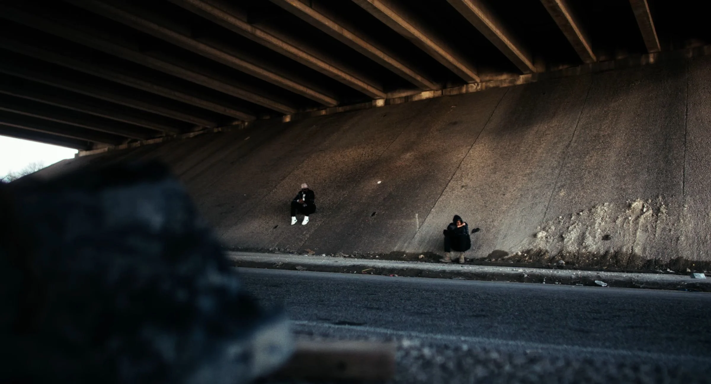 Still from INERTIA: two people sit apart against the sloped concrete wall beneath a highway overpass, a blurred rock in the foreground.