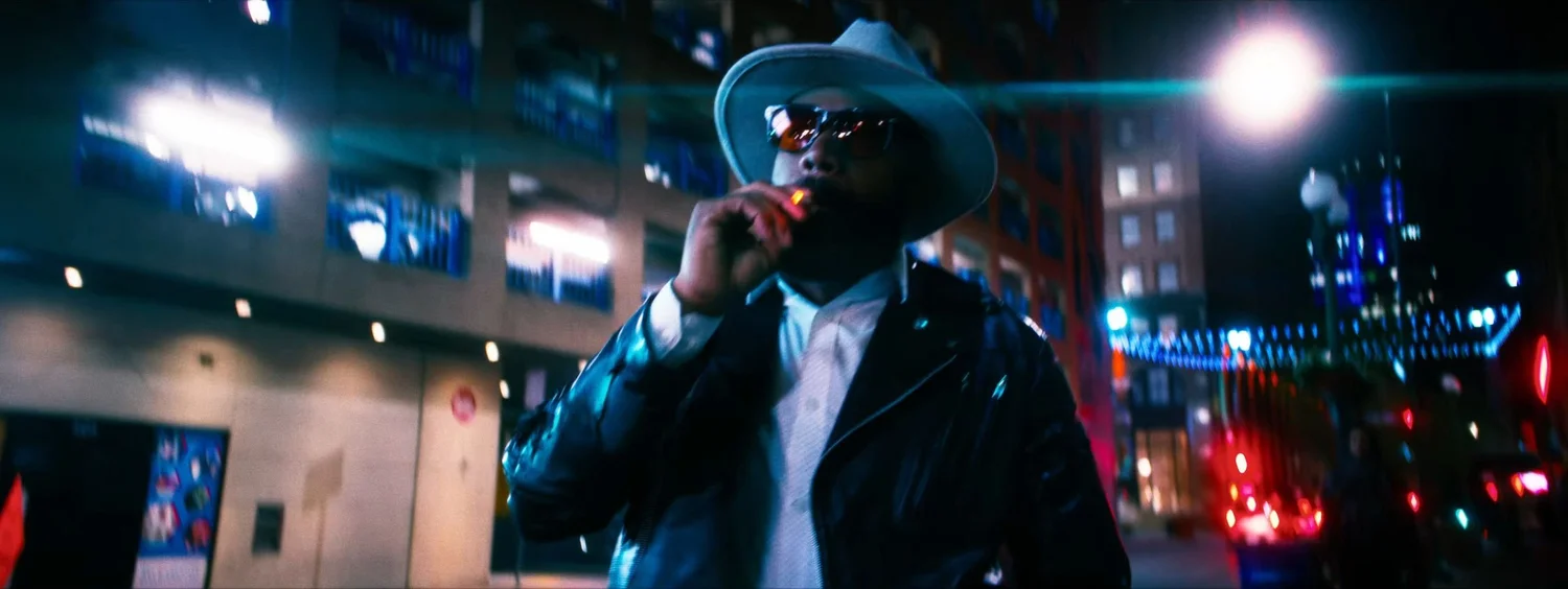 Still from INERTIA: a man in a light fedora, sunglasses and a black leather jacket smokes a cigarette on a city street at night.