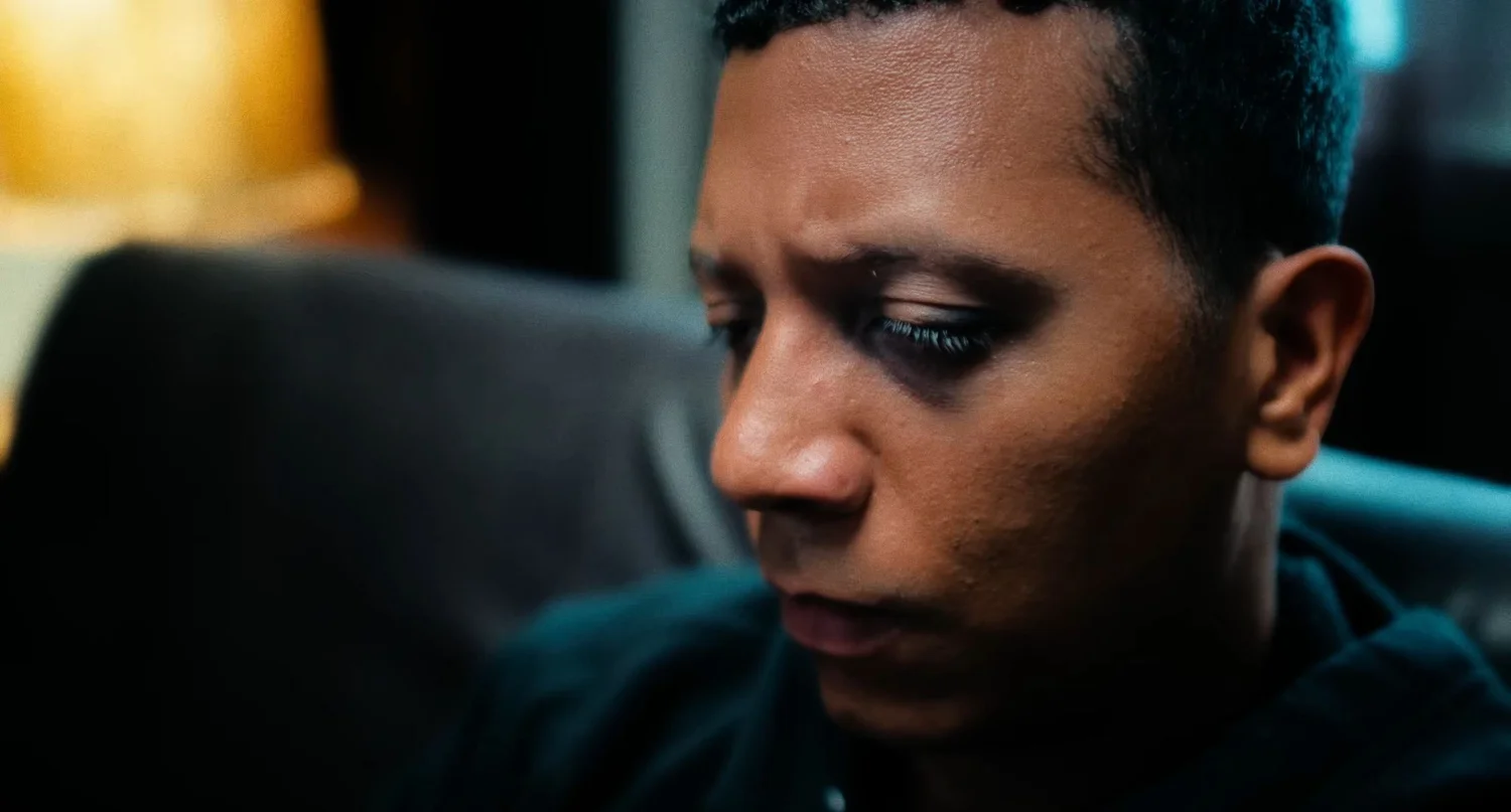 Still from INERTIA: a close view of a man with short dark hair looking down, a warm lamp glowing out of focus behind him.