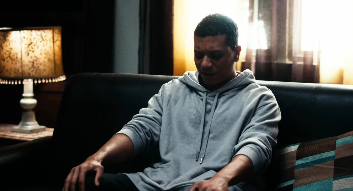Still from INERTIA: a man in a gray hoodie sits on a black leather couch looking down, a patterned table lamp and a sunlit curtained window behind him.