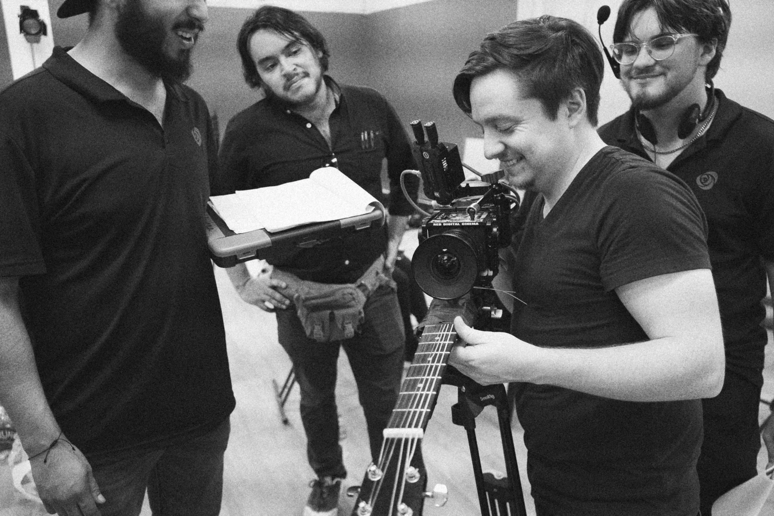 Behind the scenes on INERTIA: four crew members laugh together around a camera on a tripod, one holding an open binder and another holding the neck of a guitar.