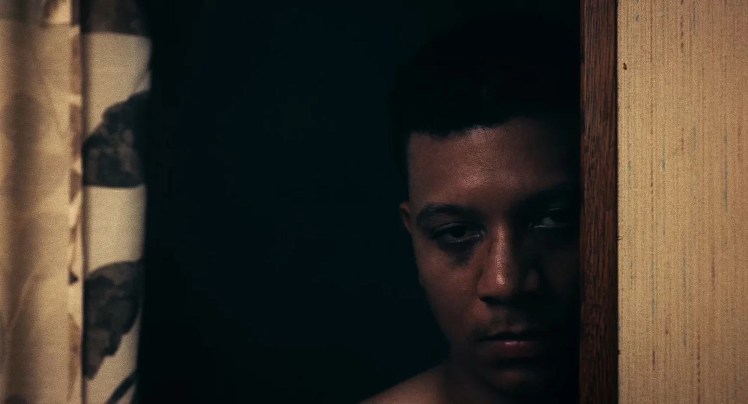 Still from INERTIA: a man's half lit face looks out from a dark doorway beside a wooden door frame, a patterned curtain at the left edge.