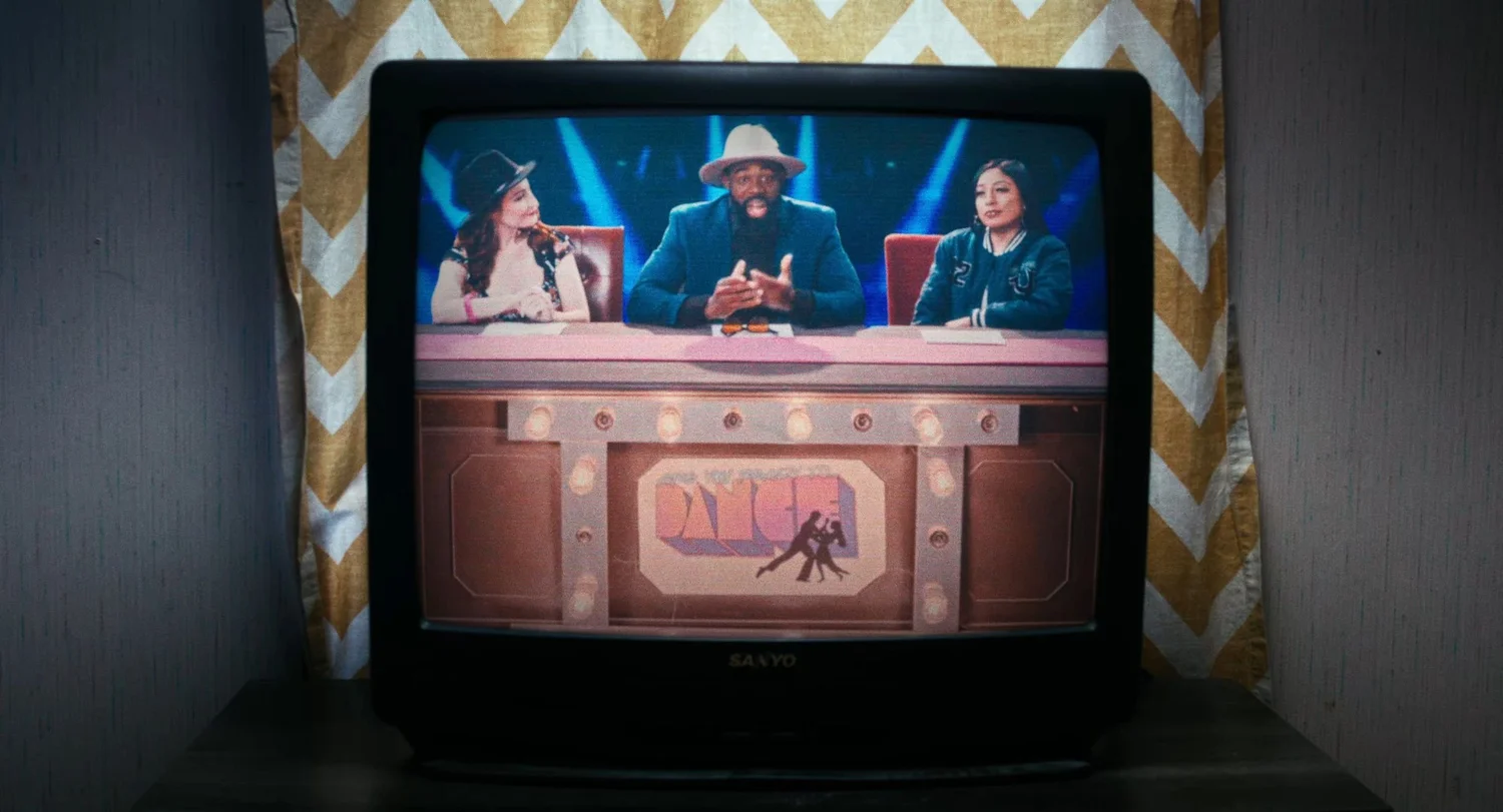 Still from INERTIA: an old television set shows three people at a pink judging desk under blue stage lights.