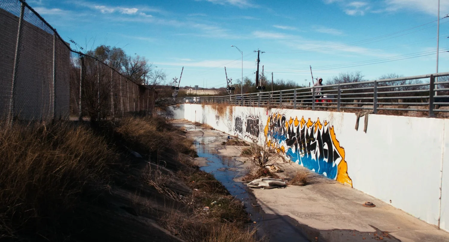 Still from INERTIA: a concrete drainage channel with shallow water and a graffiti covered white wall runs beneath a roadside railing where a person in a hooded sweatshirt stands under a blue sky.