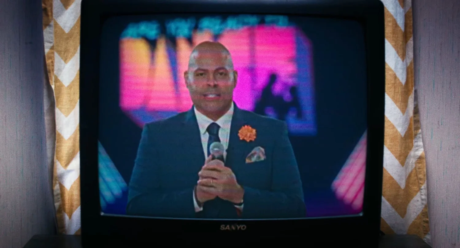 Still from INERTIA: an old television set shows a bald man in a navy suit holding a microphone in front of a bright pink stage screen, with chevron curtains behind the set.