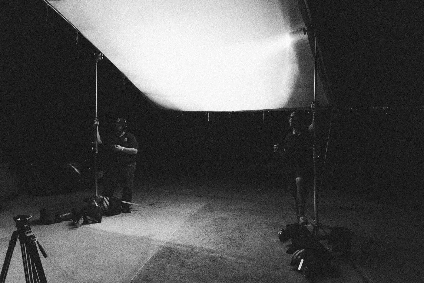 Behind the scenes on INERTIA: a large overhead silk glows above a dark driveway where crew set up lights at night.
