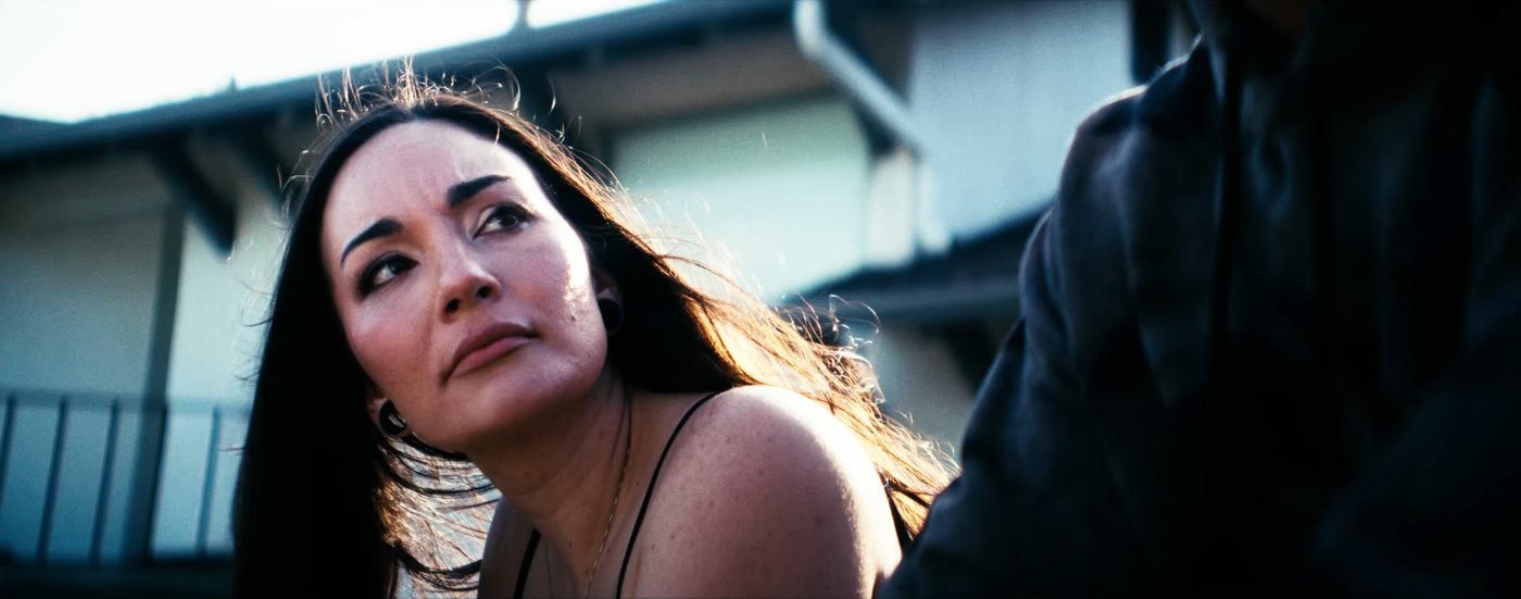 Still from INERTIA: a woman with long dark hair looks up and to the side in bright backlight, a figure in a dark jacket in the foreground at the right.