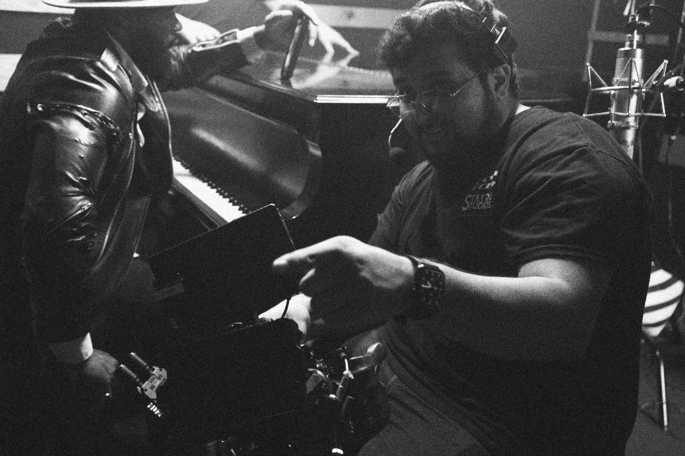 Behind the scenes on INERTIA: a crew member gestures while talking with an actor in a leather jacket seated at a piano.