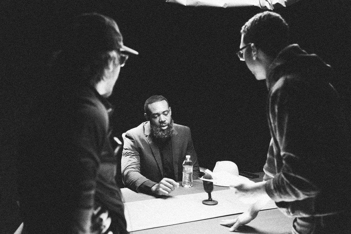 Behind the scenes on INERTIA: two crew members lean in to talk with an actor in a blue suit seated at a pink table.