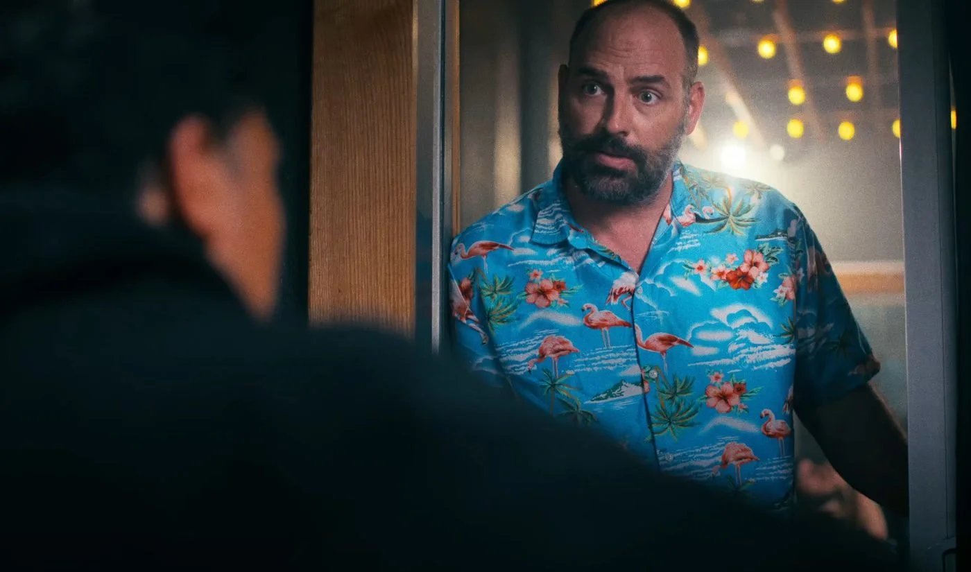 Still from INERTIA: a man in a flamingo print shirt stands in a doorway with warm string lights behind him, seen past the dark shoulder of a man in the foreground.
