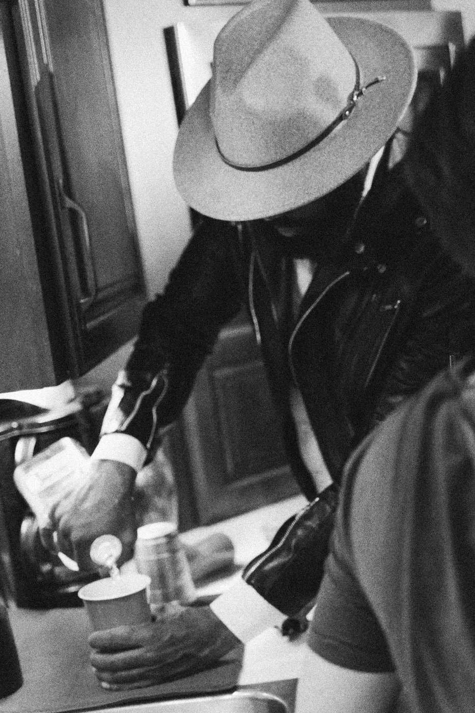 Behind the scenes on INERTIA: a man in a wide brimmed hat pouring a drink into a red cup at a kitchen counter.