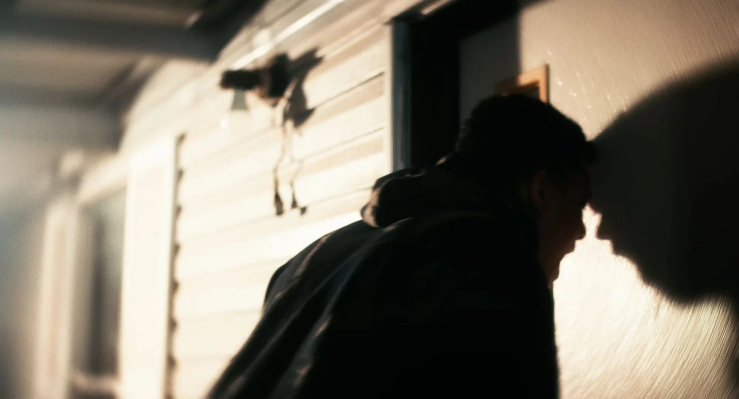 Still from INERTIA: a man in a dark hooded jacket leans his forehead against the wall beside a door in harsh light, his shadow cast on the wall.