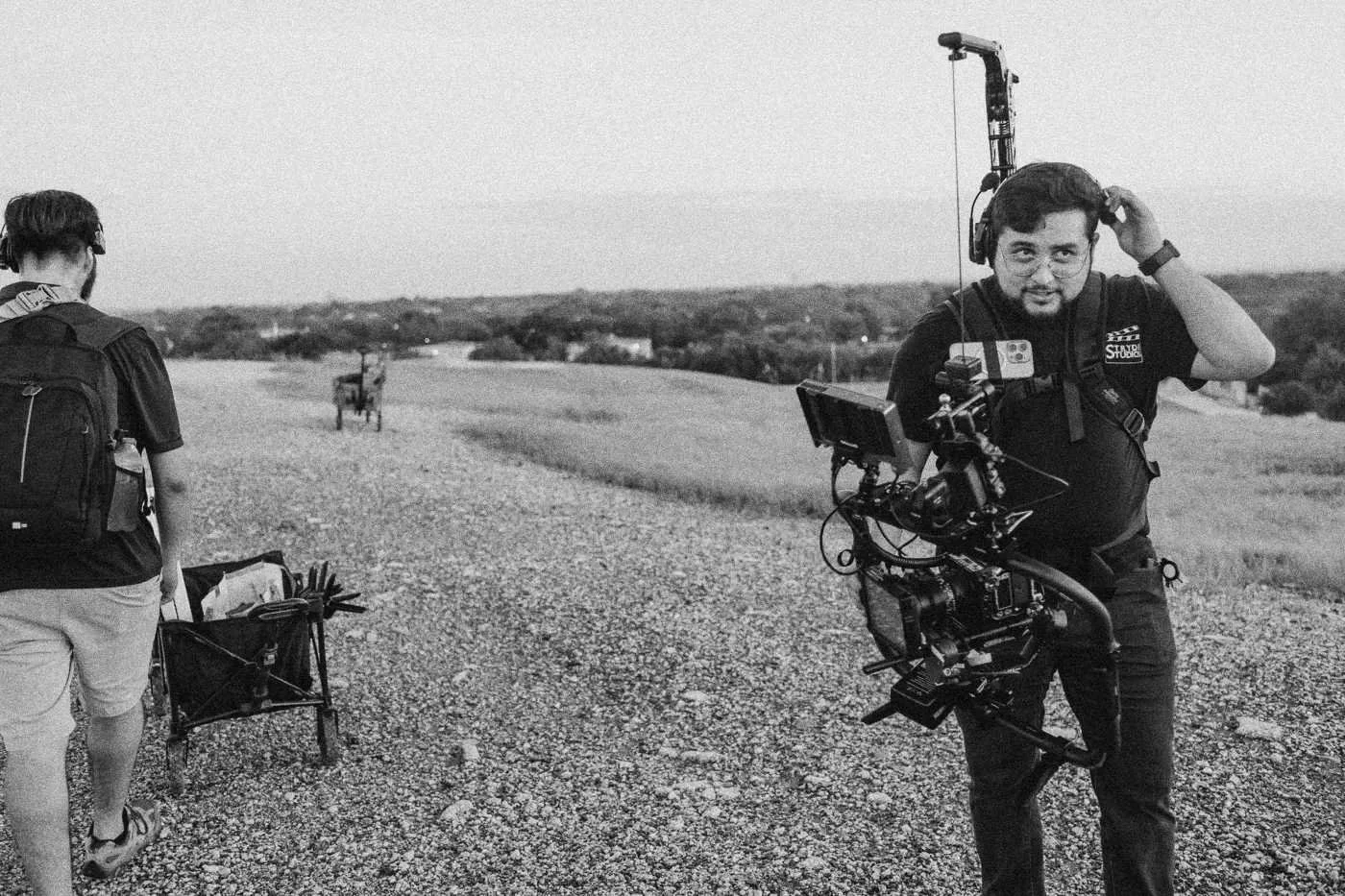 Behind the scenes on INERTIA: a camera operator with a gimbal rig adjusts his cap on a gravel hilltop, crew carrying gear behind.