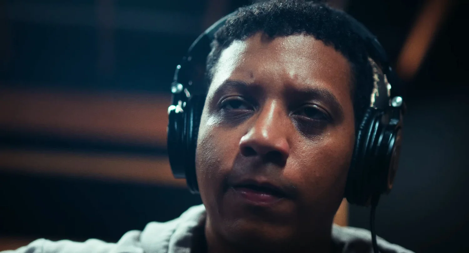 Still from INERTIA: a close up of a man wearing black studio headphones looking just past the camera, blurred warm stripes of light behind him.