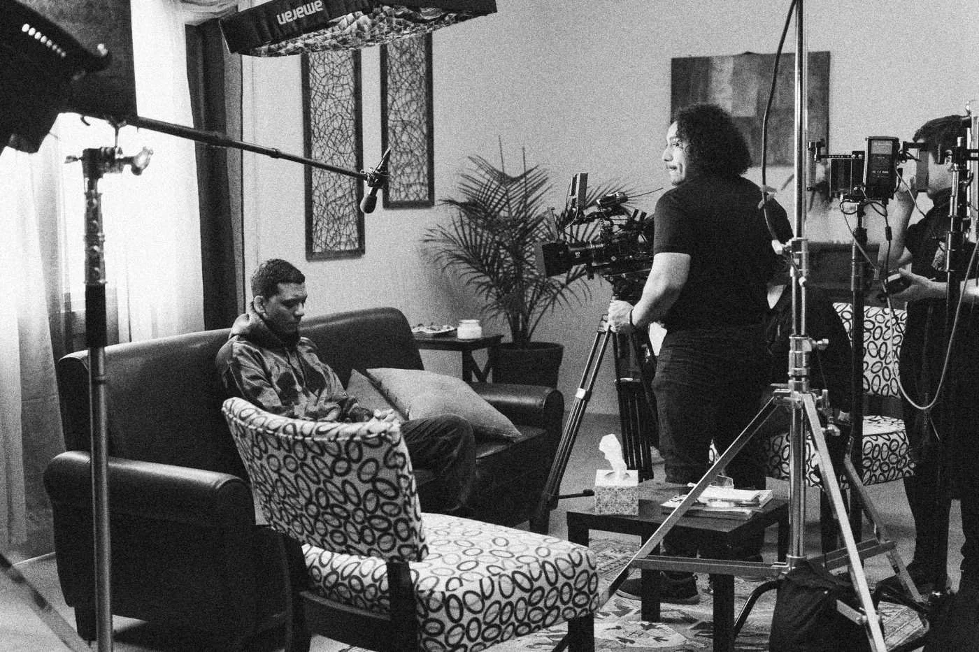 Behind the scenes on INERTIA: wide view of the living room set with a seated actor surrounded by camera, lights and crew.