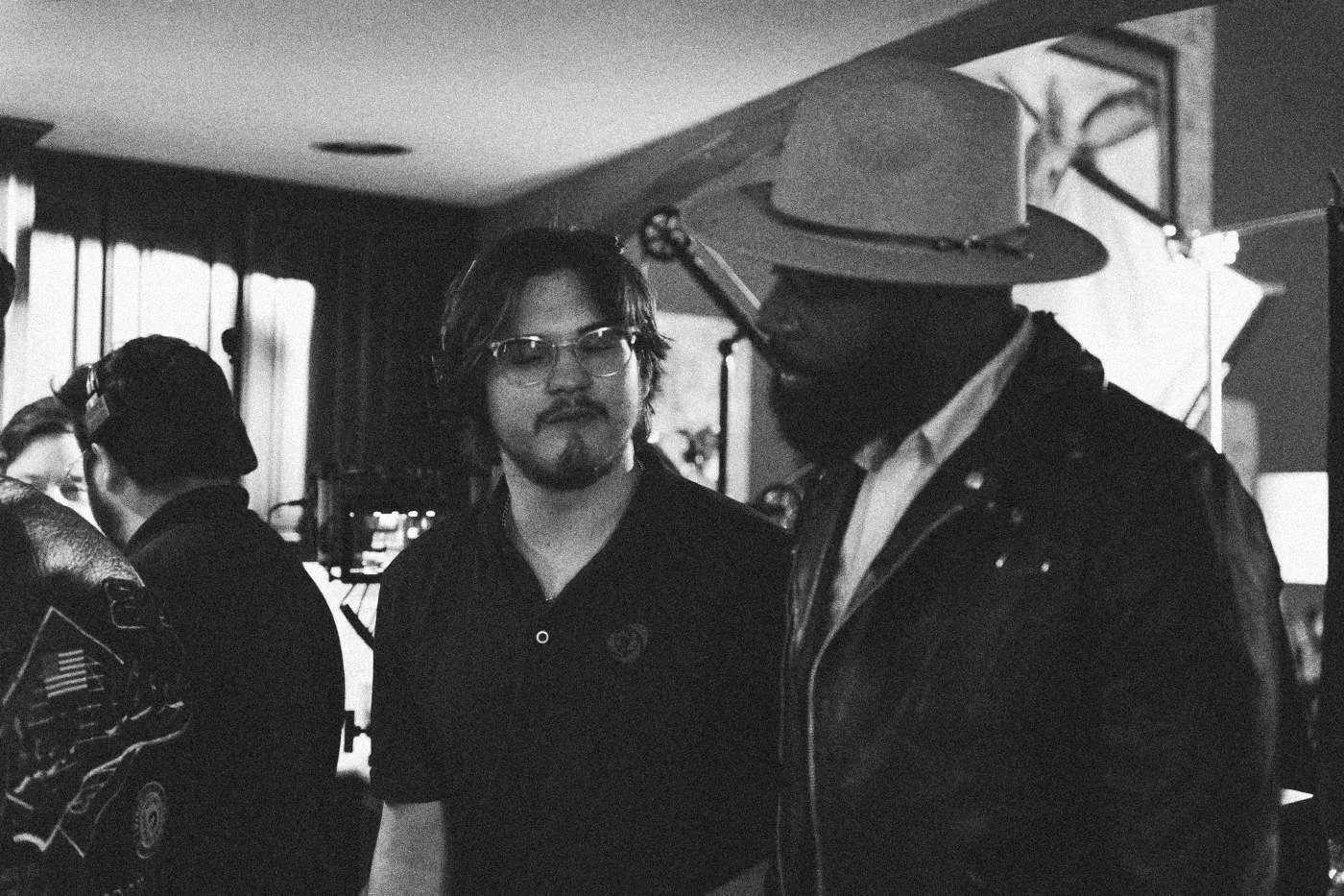 Behind the scenes on INERTIA: a crew member in glasses talks with an actor in a wide brimmed hat and leather jacket.