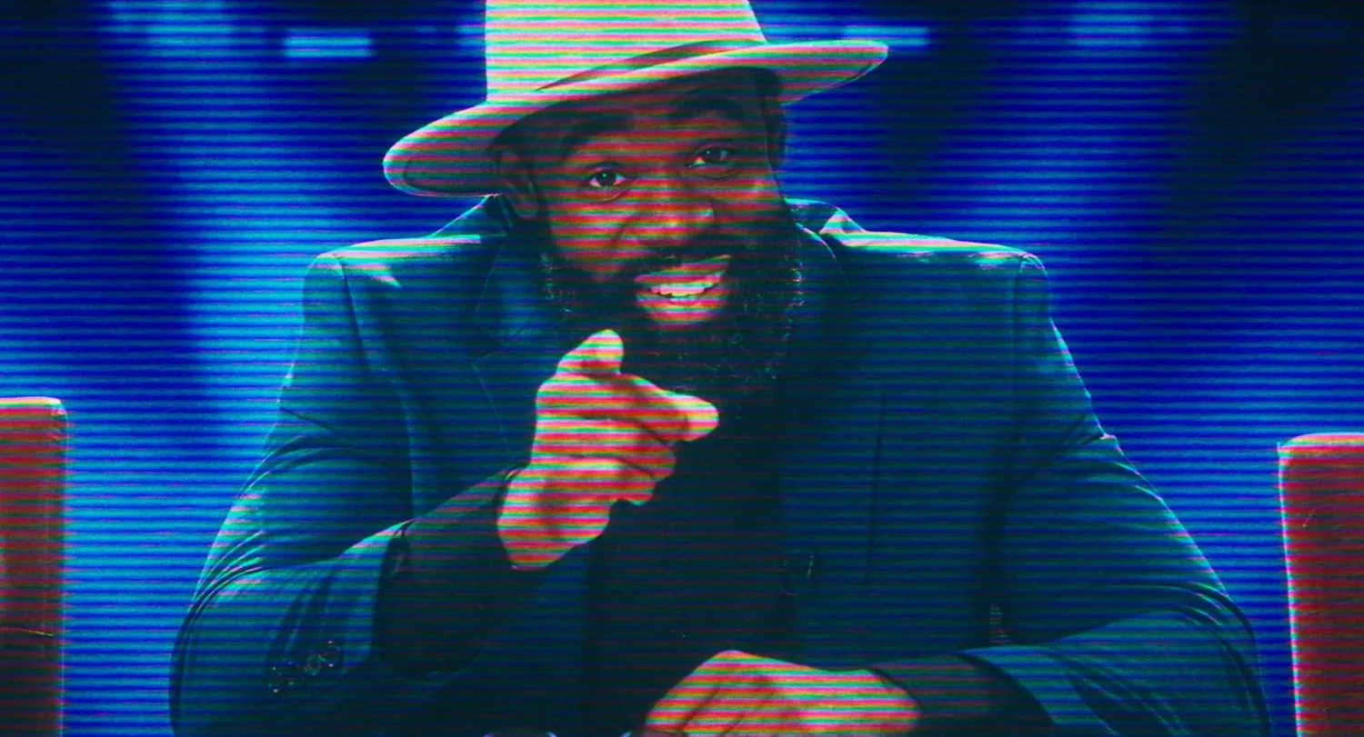 Still from INERTIA: a bearded man in a wide brimmed hat and dark blazer grins and points at the camera, tinted blue with television scan lines.
