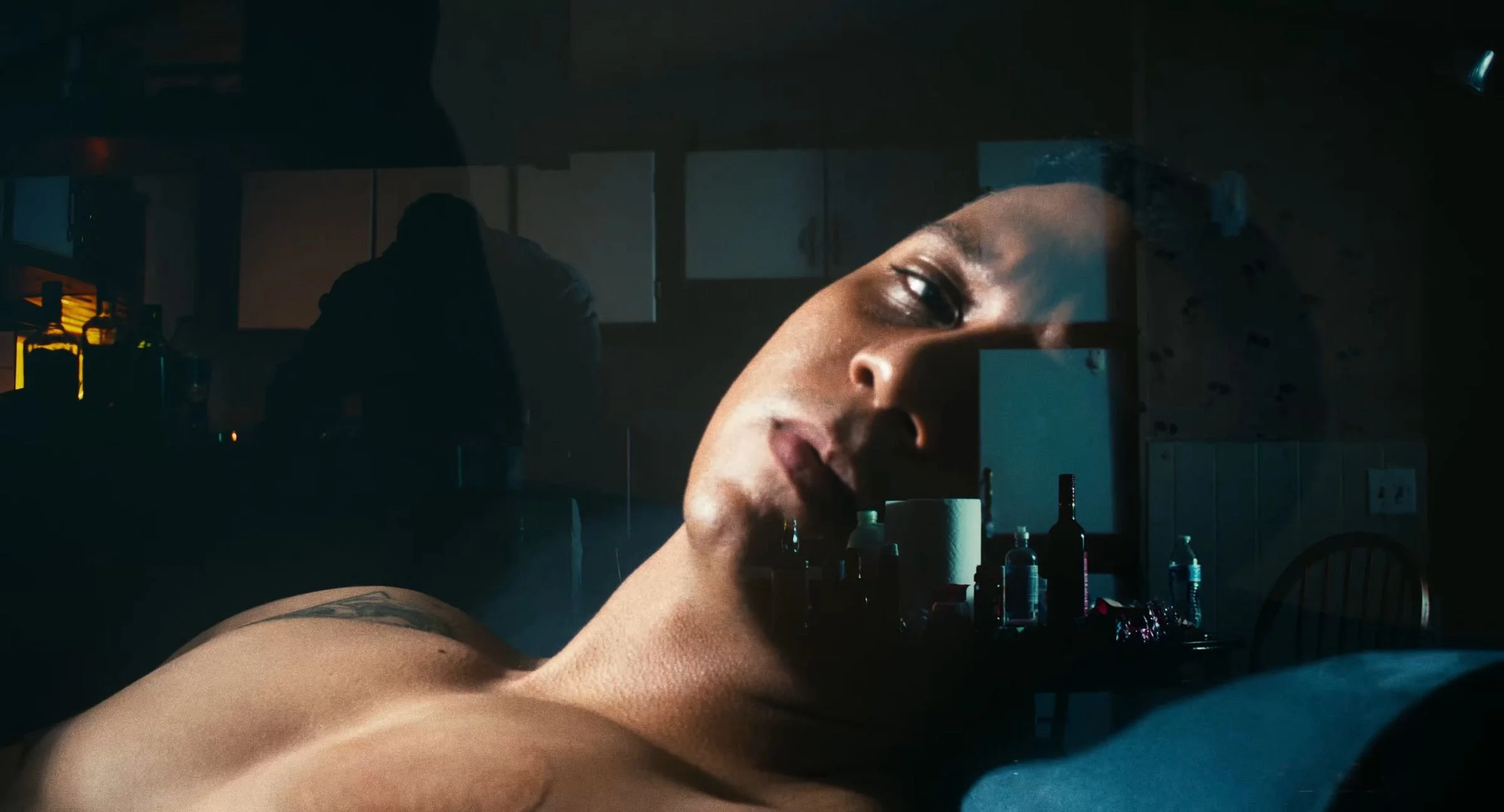 Still from INERTIA: a double exposure of a shirtless man lying back with his eyes half closed, laid over a dim kitchen where a silhouetted figure stands at a counter and bottles crowd a table.