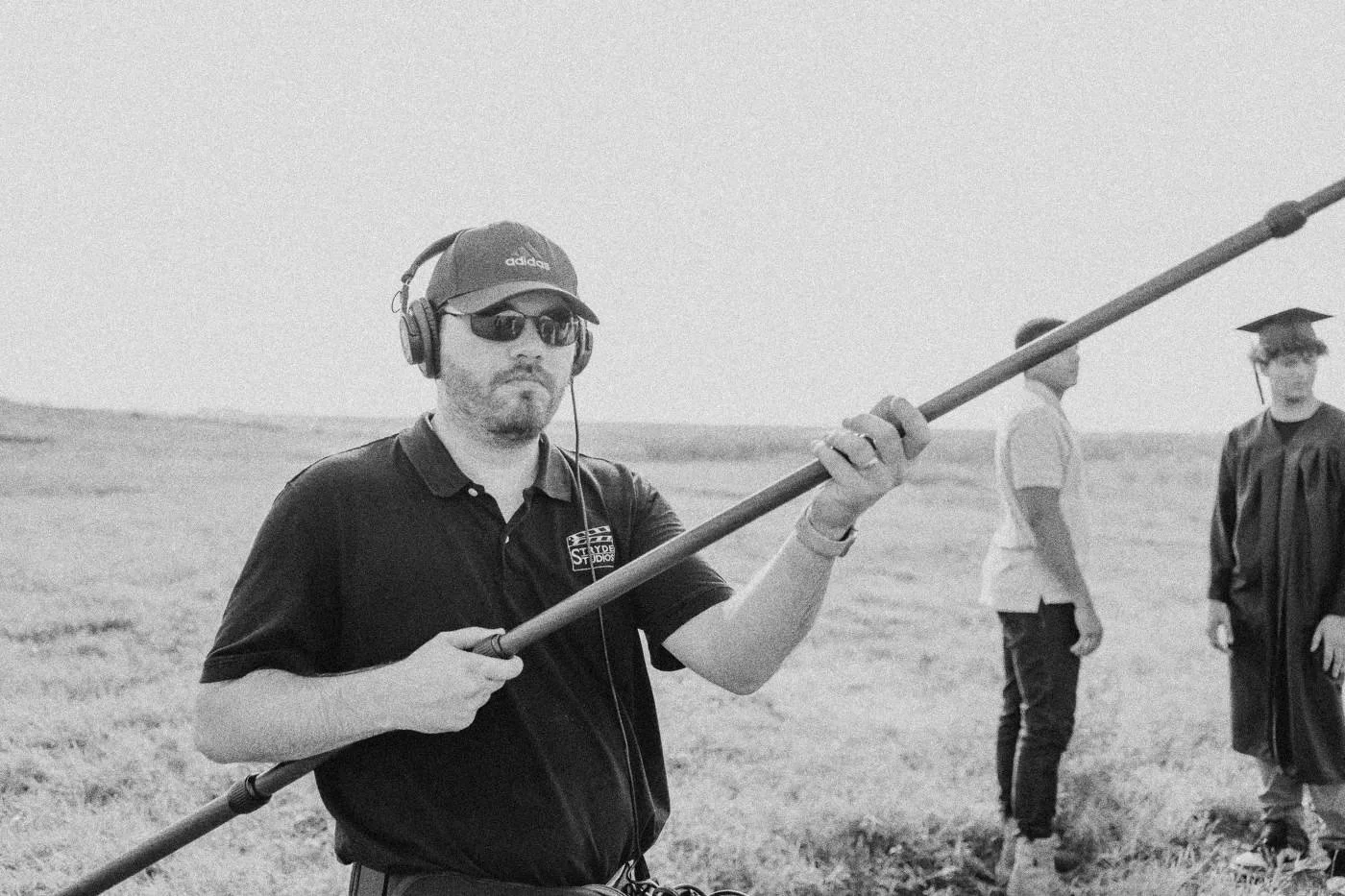 Behind the scenes on INERTIA: a boom operator in sunglasses swings the pole in a field while cast in graduation gowns wait behind.