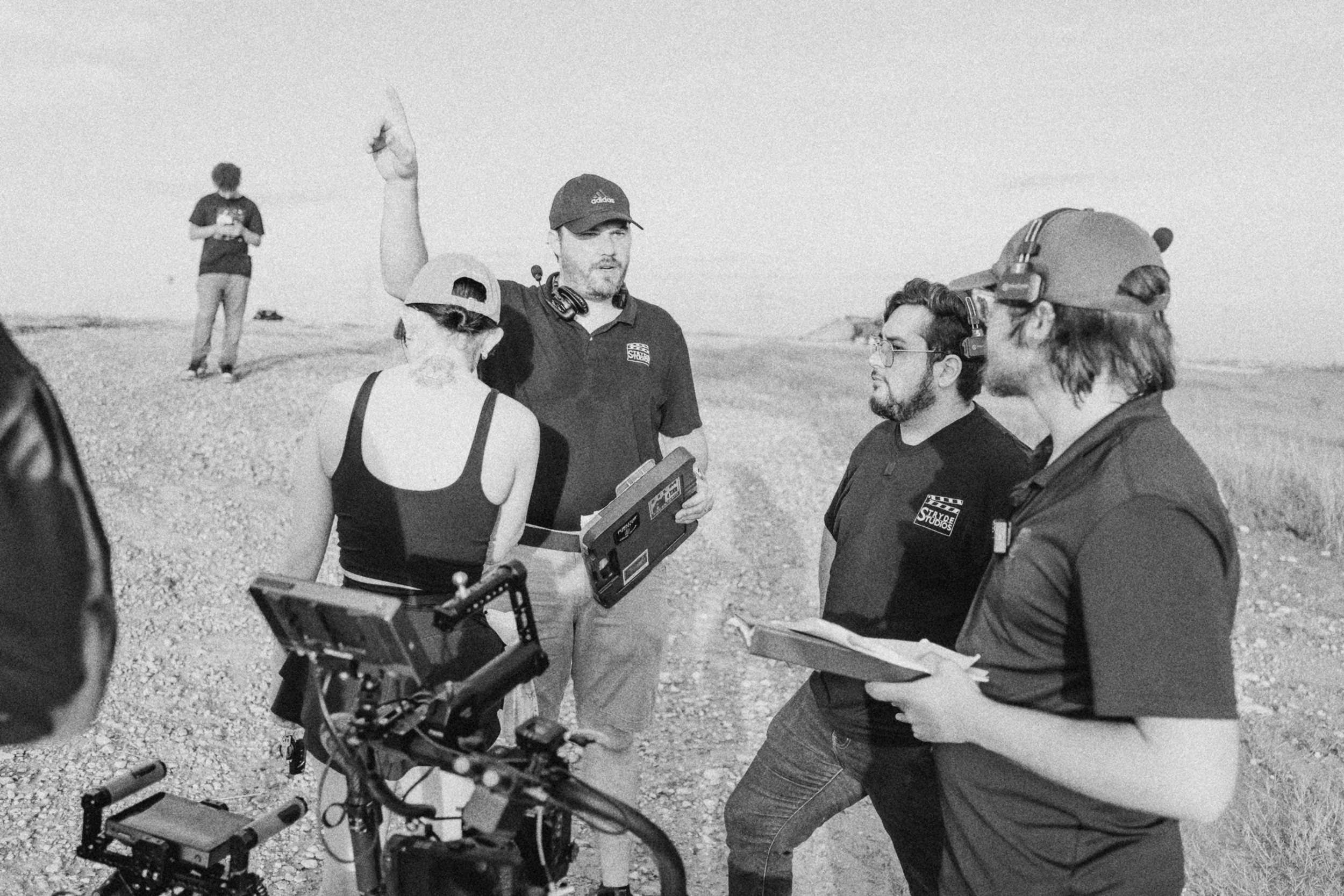 Behind the scenes on INERTIA: on a gravel hilltop a crew member in a cap points to the sky while the others gathered around a camera rig listen, one holding a folder of pages.