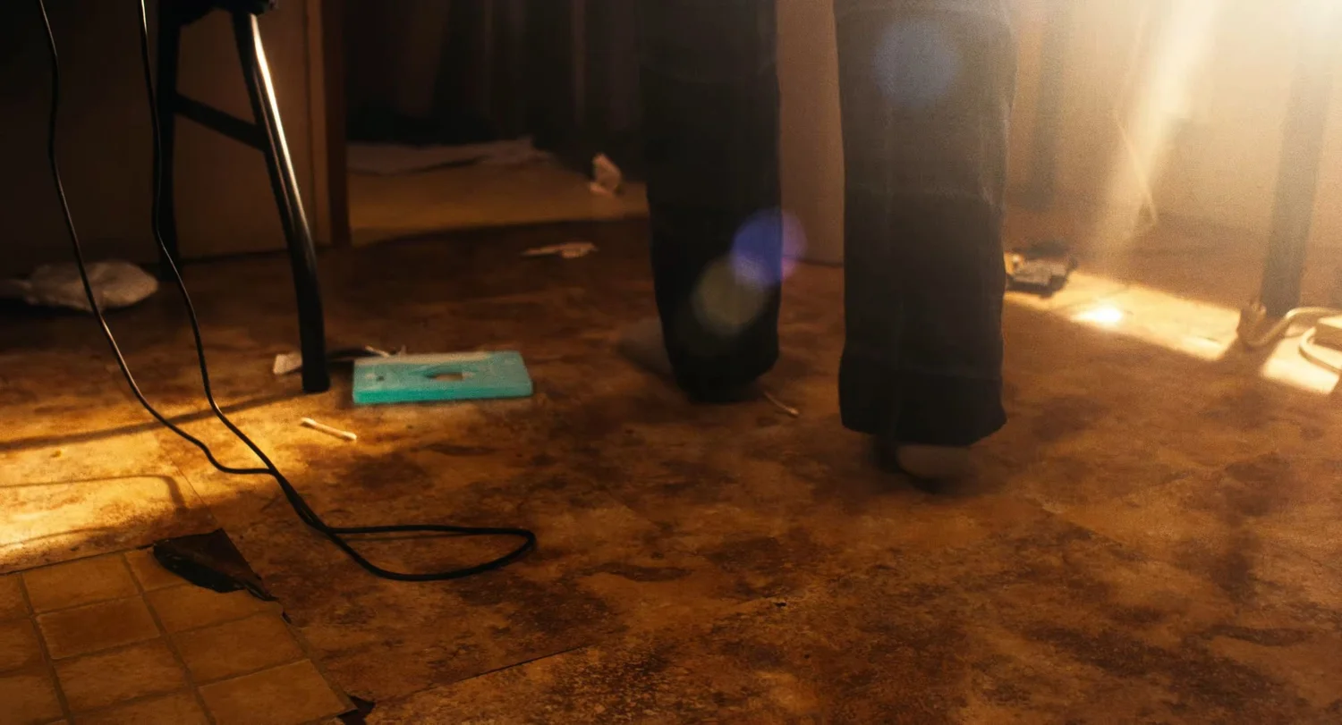 Still from INERTIA: a low view of a littered floor in warm sunlight, with the legs of a person in dark trousers, a teal cassette case and a loose black cable.
