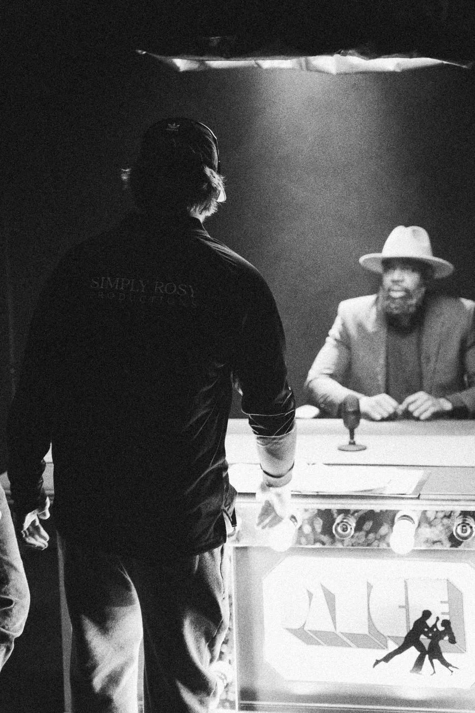 Behind the scenes on INERTIA: a crew member walking toward a lit desk where an actor in a hat sits.