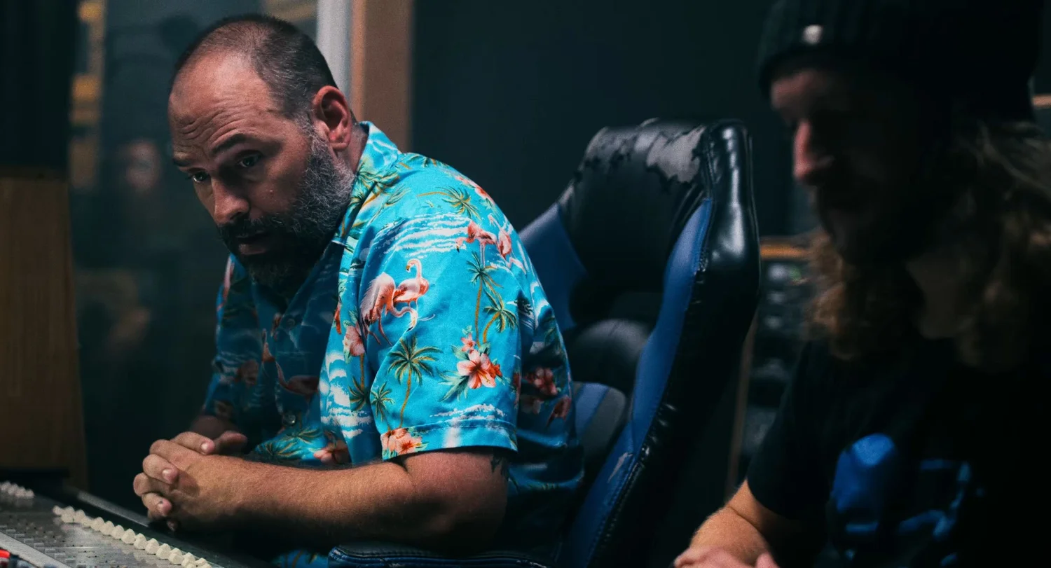Still from INERTIA: a man in a flamingo print shirt leans on a mixing console beside an empty leather chair while a long haired man in a beanie sits to the right.