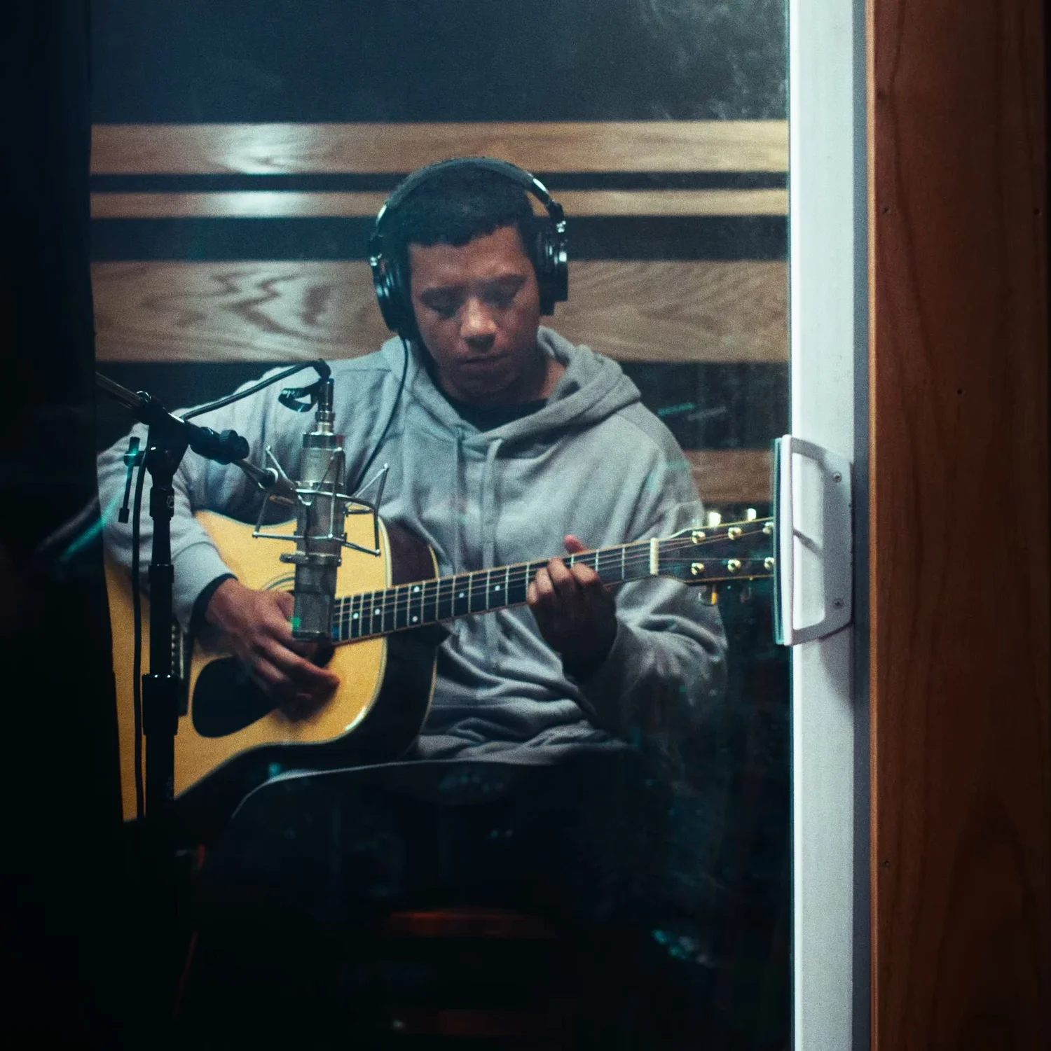 Still from INERTIA: a man in headphones and a gray hooded sweatshirt plays an acoustic guitar into a studio microphone, seen through the glass of a booth door framed by darkness.