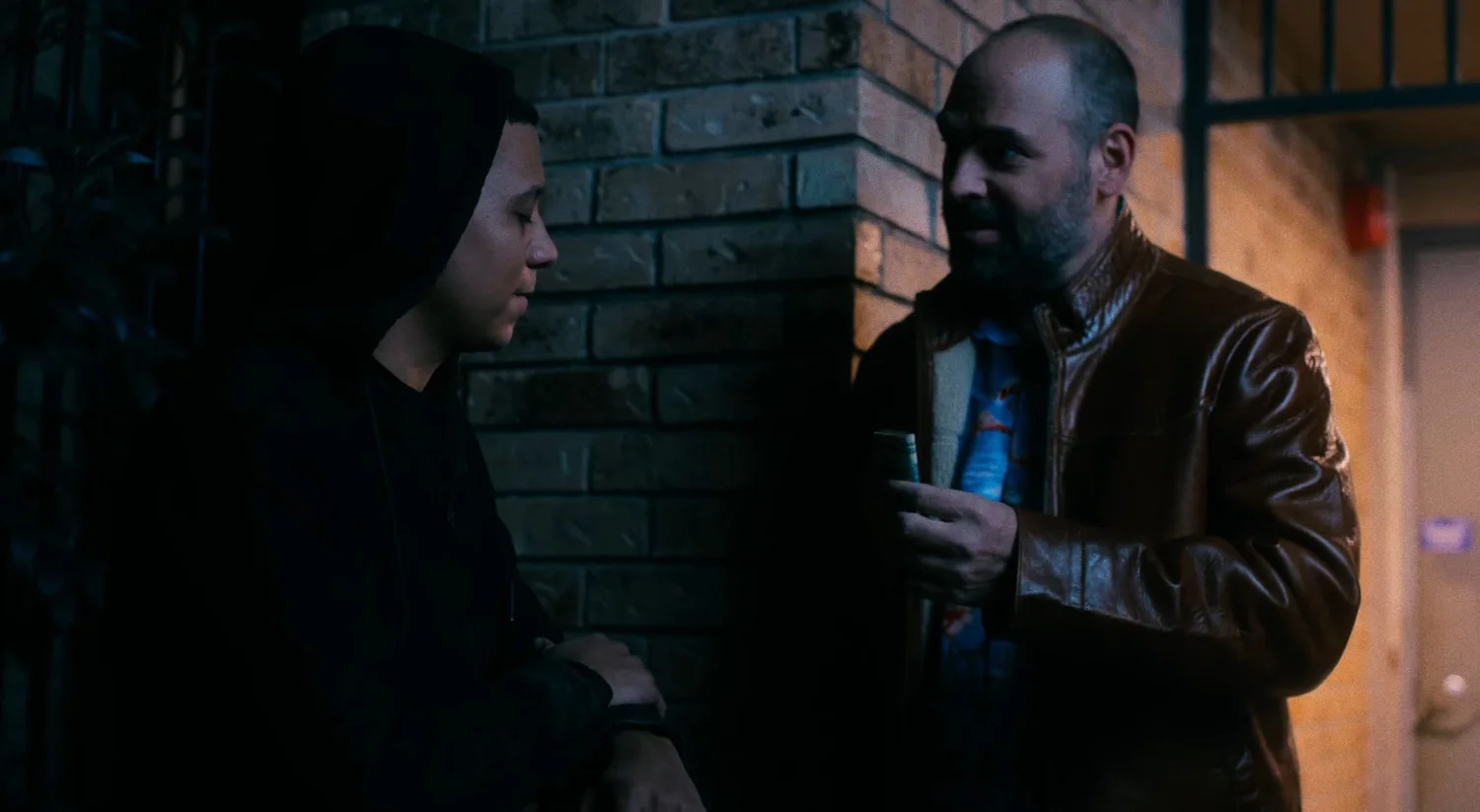 Still from INERTIA: at night beside a brick wall, a young man in a black hoodie faces a bearded man in a brown leather jacket who holds a small roll of bills.