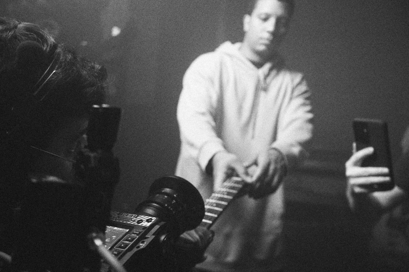 Behind the scenes on INERTIA: an actor in a hoodie holds a guitar while crew film him, one holding up a phone.