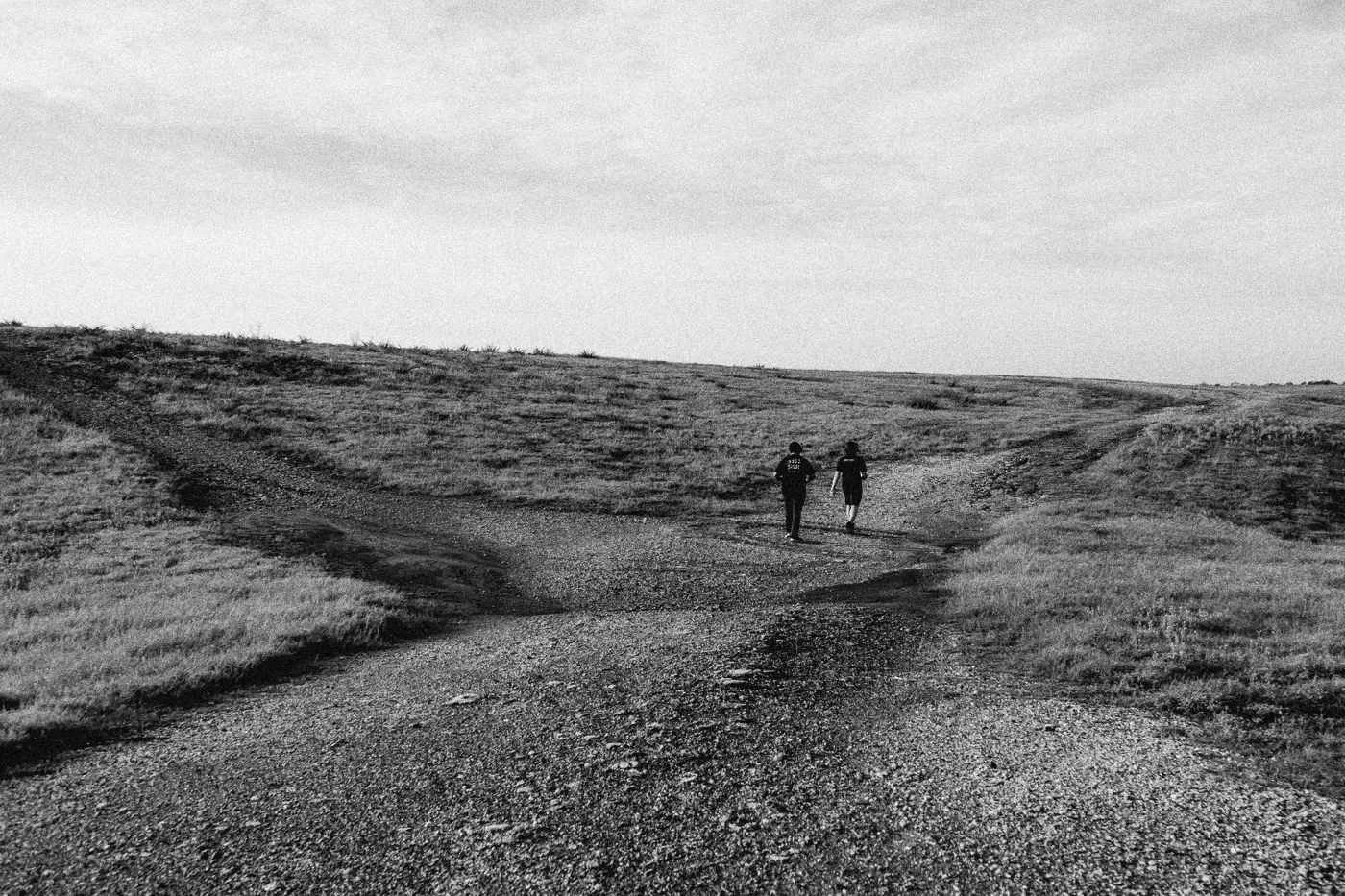 Behind the scenes on INERTIA: two figures walk down a dirt road between grassy slopes under a wide sky.