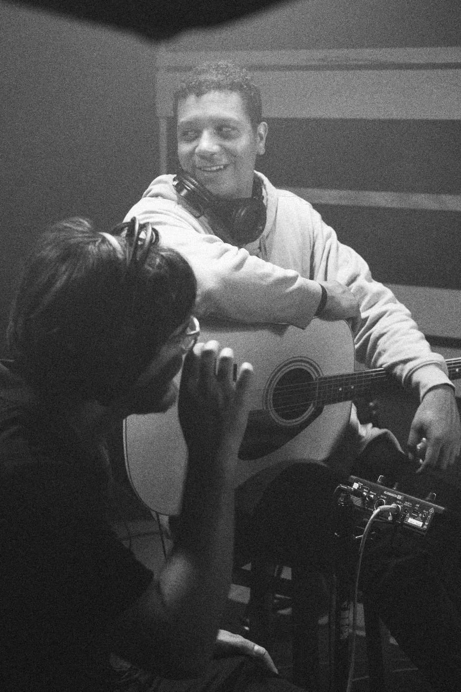 Behind the scenes on INERTIA: an actor with an acoustic guitar smiling, a crew member in the foreground.