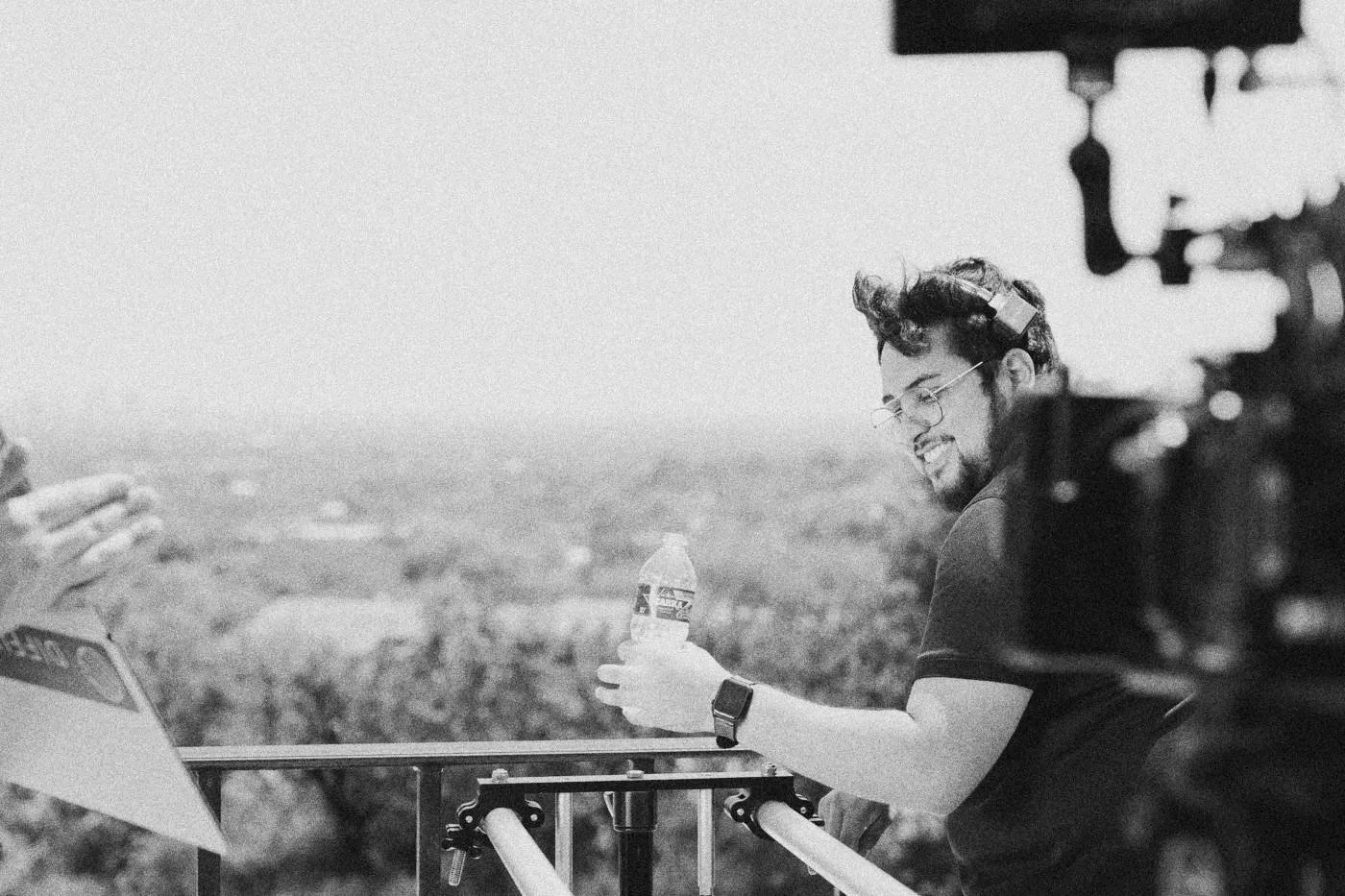 Behind the scenes on INERTIA: a camera operator smiles while holding a water bottle on a hilltop deck, trees and a city haze behind.
