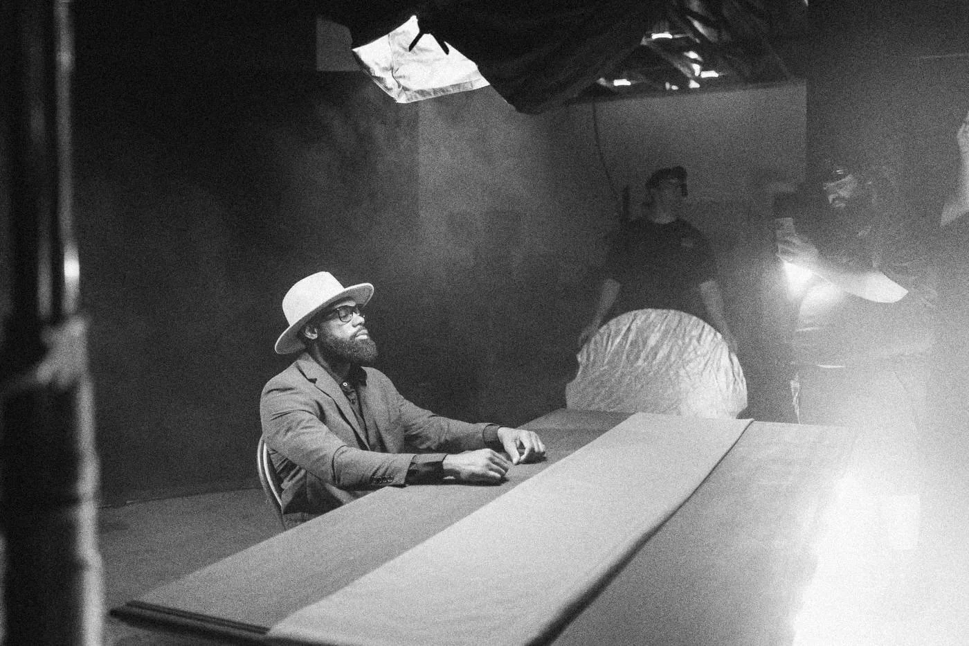 Behind the scenes on INERTIA: an actor in a hat and blue suit sits at a pink table as haze drifts and crew work behind him.
