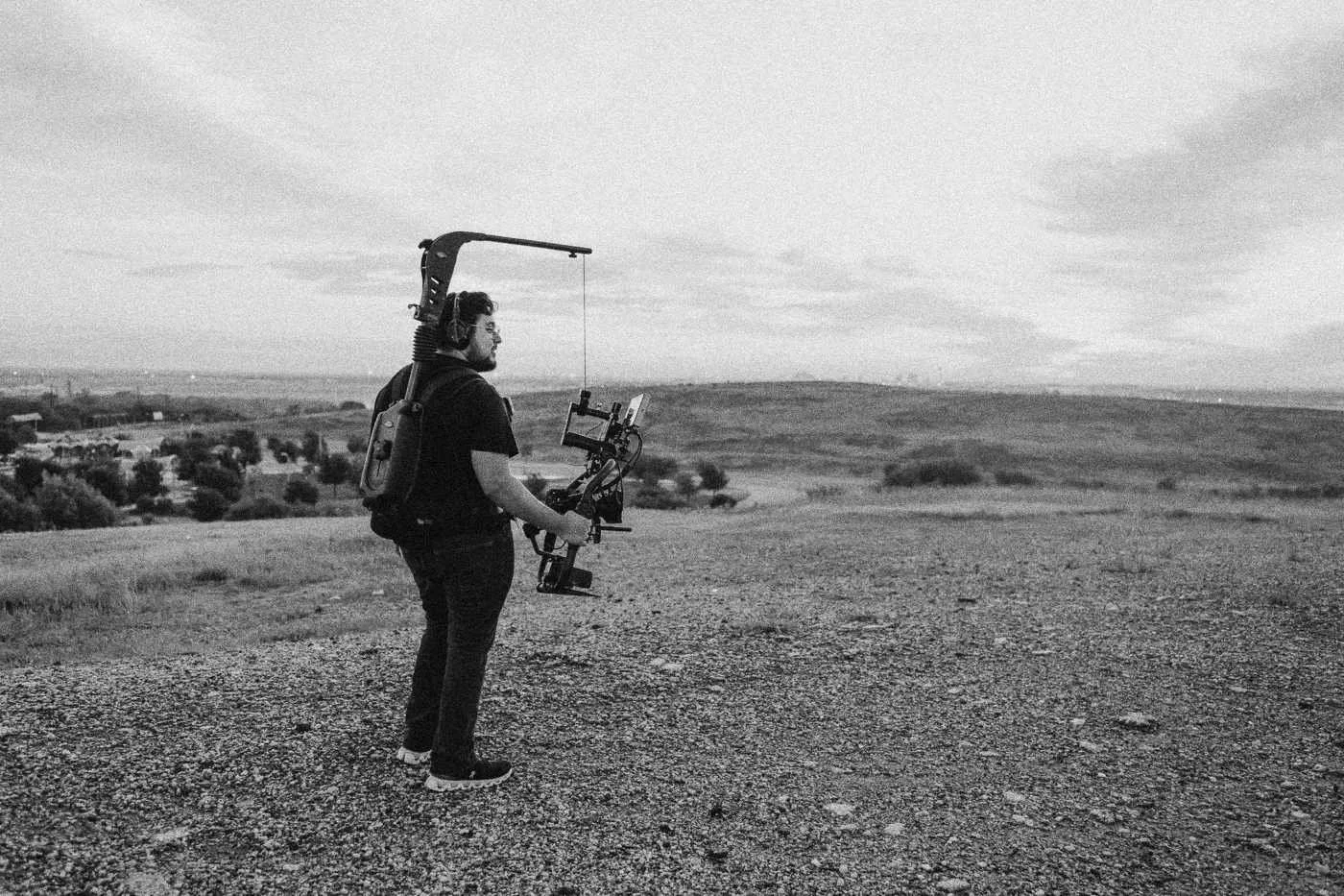 Behind the scenes on INERTIA: a camera operator with a gimbal stands on a gravel hilltop against a sunset sky.