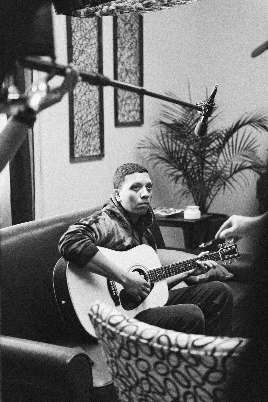 Behind the scenes on INERTIA: a man seated on a couch playing an acoustic guitar under a boom microphone.