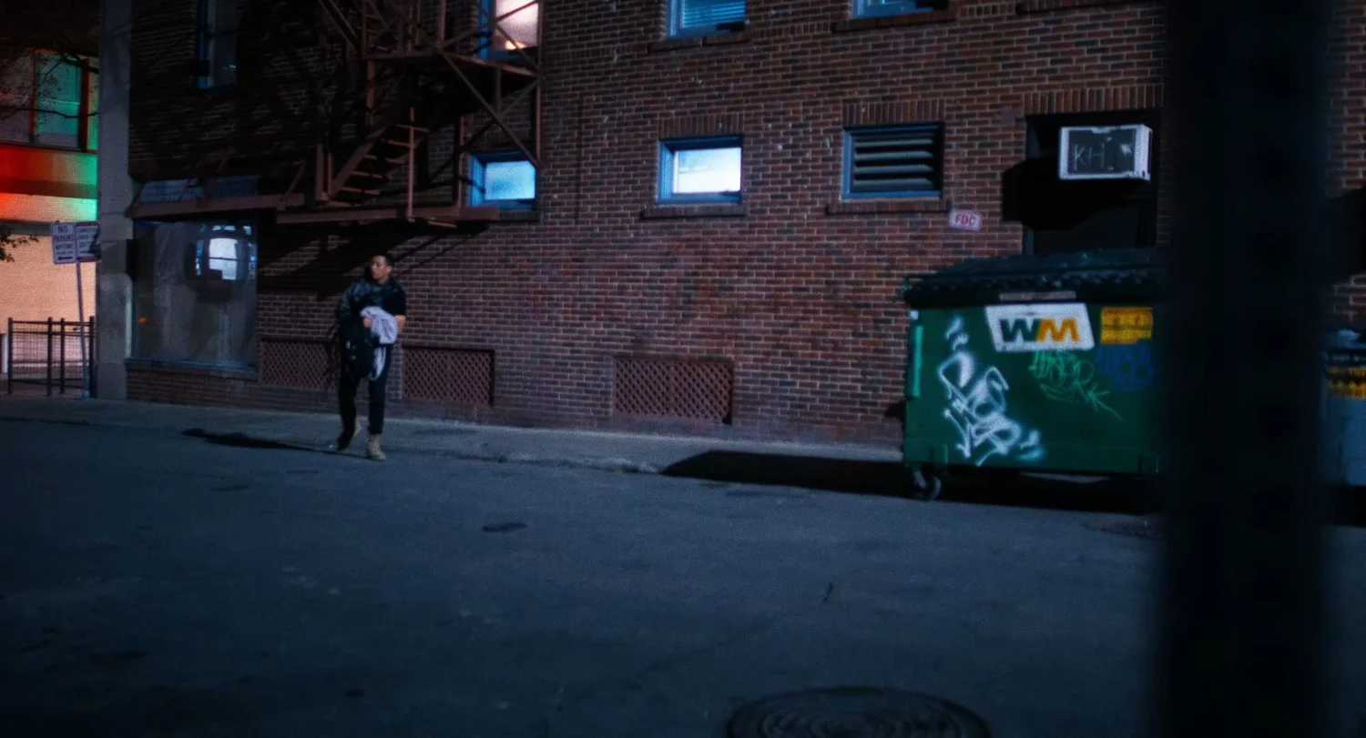 Still from INERTIA: a man in dark clothes carries a white cloth along a brick building at night, past a fire escape and a green dumpster covered in graffiti.