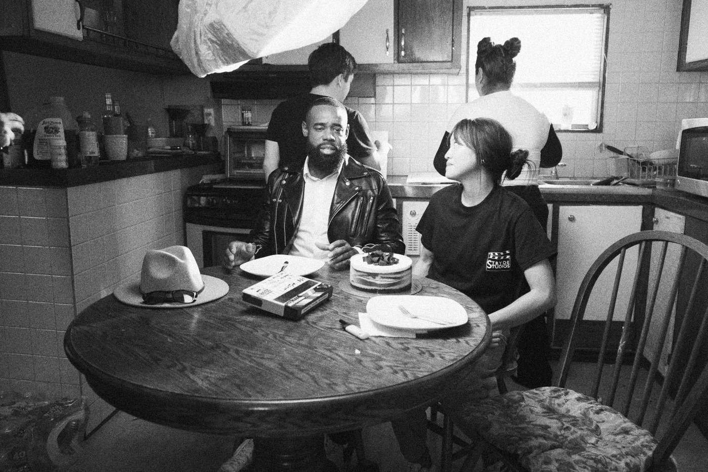 Behind the scenes on INERTIA: an actor in a leather jacket and an actress sit at a round kitchen table set with plates, a hat on the table.