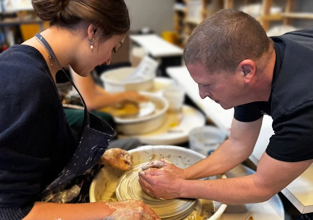 A teacher guiding a student at the pottery wheel